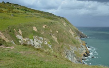 Kara bulutlar tepemizde belirdikçe dalgalar sarp kayalıklara çarpar. Yeşillik kayalıkları süsler ve doğal harikalarla dolu dramatik bir kıyı manzarası yaratır..