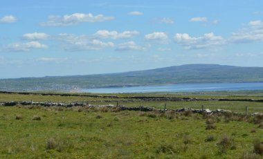 Yeşil yemyeşil tepeler araziye yayılarak huzurlu bir su kütlesine ulaşıyorlar. Yumuşak bulutlar dalgalı mavi gökyüzünde sürükleniyor, İrlanda kırsalında güneşli bir günün özünü yakalıyor..