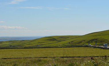 Geniş yeşil tepeler yavaşça yükselir, açık mavi gökyüzüne karşı çarpıcı bir kontrastlık yaratır. Uzak kıyı şeridi bu huzurlu kırsal bölgeye güzellik katıyor. Keşif ve yansımayı davet ediyor..