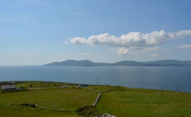 Yuvarlanan yeşil tepeler sakin bir denize doğru uzanıyor, uzak dağlar canlı mavi bir gökyüzü altında kabarık bulutlarla kaplı. Huzurlu bir sahne başlıyor, rahatlatıcı..