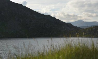 Sakin sular etrafındaki tepeleri yansıtıyor. Yumuşak bulutlar yukarı doğru sürükleniyor. Bereketli yeşil çimenler rüzgarda hafifçe sallanıyor, bu doğal ortamda huzurlu bir atmosfer yaratıyor..