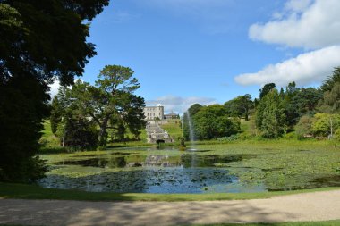 Bereketli yeşillik, bir çeşmenin zarifçe su fışkırttığı sakin bir göleti çevrelemektedir. Güneşli bir öğleden sonra canlı ağaçlar tarafından çerçevelenmiş, arka planda ihtişamlı bir klasik malikane duruyor..