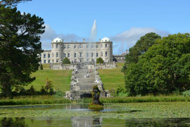Heybetli bir malikane heybetli bir tepenin üzerinde duruyor, serin bir gölete doğru yükselen basamaklar. İnsanlar güneşli bir günde yemyeşil bahçelerde gezinir, manzaranın tadını çıkarırlar..