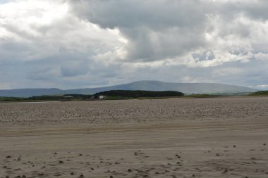 Açık kumlu sahil, bulutlar ve güneş altında geniş bir alana yayılır. Uzak dağlar nazikçe yükselir, doğa severlerin keşfetmesi için huzurlu bir zemin oluşturur..