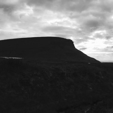 Bir Stark tepesi alacakaranlıkta bulutlu bir gökyüzüne çarpıcı bir şekilde yükselir. Gölgeler arazide oynar, dingin manzarada bir gizem ve entrika hissi yaratır..