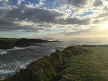 Güneş ufukta batarken dalgalar yavaşça sahili öpüyor. Yumuşak bulutlar ve dingin bir okyanus ile dolu huzurlu bir sahneyi çerçeveleyen yemyeşil uçurumlar yükselir..