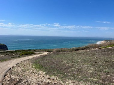 Dolambaçlı bir toprak yol, mavi okyanusun gökyüzüyle buluştuğu nefes kesici bir kıyı manzarasına götürür. Kıyı şeridi, yeşil tepeler ve canlı doğa tarafından çerçevelenmiş olan mesafeye kadar uzanıyor..