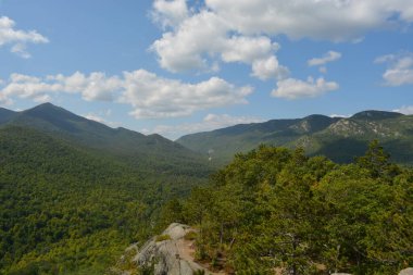 Ufuk boyunca uzanan yemyeşil dağlar, parlak mavi gökyüzünün altında kabarık bulutlarla kaplı canlı ağaçlarla çevrili. Barışçıl bir doğa sahnesi açık havanın güzelliğini gözler önüne seriyor..