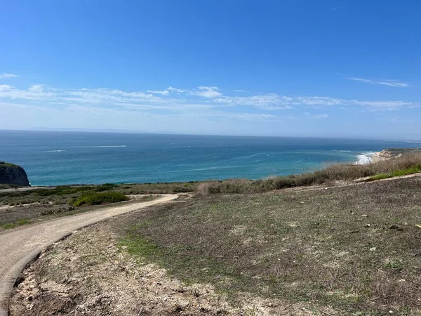 Dolambaçlı bir toprak yol, mavi okyanusun gökyüzüyle buluştuğu nefes kesici bir kıyı manzarasına götürür. Kıyı şeridi, yeşil tepeler ve canlı doğa tarafından çerçevelenmiş olan mesafeye kadar uzanıyor..