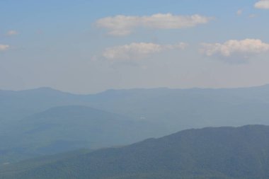 Çarpıcı mavi bir manzara katmanlı dağ sıralarının uzaklığa doğru kaybolduğunu gösteriyor. Yumuşak bulutlar tembelce tepemizde sürükleniyor, huzurlu bir günde sakin bir atmosfer yaratıyor..