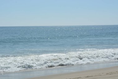 Parlak güneş, sakin okyanus suyunda parıldıyor yumuşak dalgalar kumlu kumsalda yuvarlanırken. Huzurlu bir atmosfer, kıyı şeridinde rahatlamaya ve keyif almaya davet ediyor..