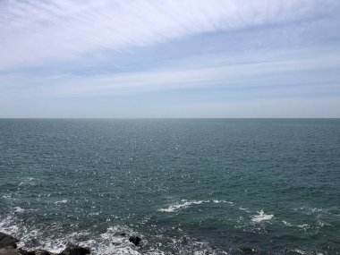 Durgun mavi okyanus, kayalık kıyı şeridini hafifçe okşayarak durmaksızın uzanıyor. Yumuşak bulutlar parlak bir gökyüzünde sürükleniyor, sakin bir atmosfer yaratıyor tefekkür için mükemmel.