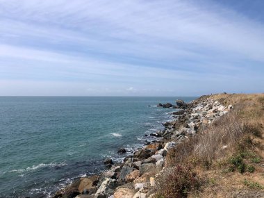 Dalgalar engebeli kıyı şeridine nazikçe yaklaşırken kabarık bulutlar gökyüzünde sürükleniyor. Rüzgarda sallanan çimenler, sahil boyunca huzurlu bir atmosfer yaratıyor..