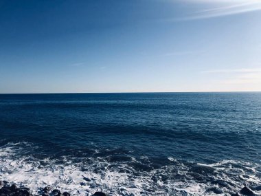 Işıl ışıl mavi sular ufka doğru uzanırken, yumuşak dalgalar engebeli kıyı şeridine hafifçe çarpıyor. Sahne, huzurlu bir sahil sabahının güzelliğini ve huzurunu yakalıyor..