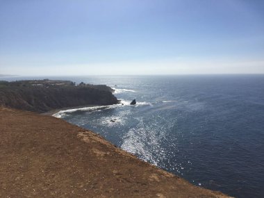 Parlak güneş ışığı, yumuşak dalgaların deniz kıyısında kayalık kayalıklarla buluştuğu sakin bir manzara çizer. Uçsuz bucaksız okyanus ufka doğru uzanıyor, huzurlu ve huzurlu bir kıyı manzarası yaratıyor..
