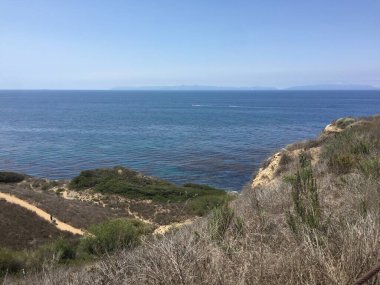 Güneşli bir sahil yolu bir uçurum boyunca esiyor, engin okyanus manzarasını ve arka plandaki uzak dağları ortaya çıkarıyor. Doğa meraklıları yürüyüş yaparken huzurlu manzaranın tadını çıkarırlar..
