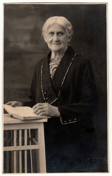 Vintage photo shows old woman with book poses in a photography studio. Photo with dark sepia tint. Black & white studio portrait.