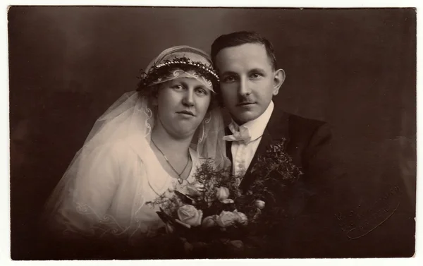 Vintage photo of newlyweds with wedding bouquet. Bride wears wedding veil headdress. Groom wears posh clothing, white bow-tie. Black & white antique studio portrait. — Stock Photo, Image