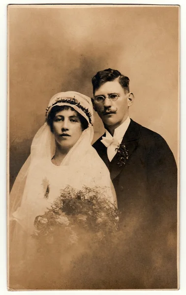 Vintage photo of newlyweds. Bride wears a veil and holds wedding bouquet. Groom wears black suit and white bow tie. Black & white antique studio portrait