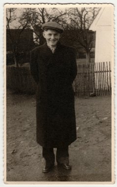 Vintage photo shows young man wears peaked cap, poses outdoors. Wooden fence and house is on background. Black & white photography.