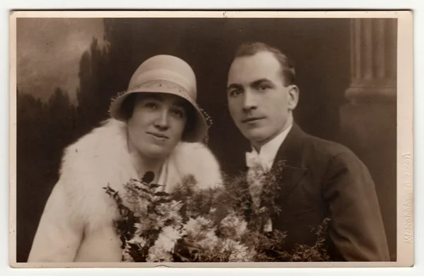 Vintage photo of newlyweds. Bride wears posh womens bowler hat and holds wedding bouquet. Groom wears black suit and white bow tie. Black & white antique studio portrait with sepia effect. — Stock Photo, Image