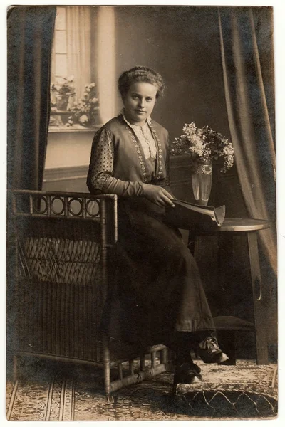 复古照片显示年轻女子坐在柳条扶手椅上，在摄影棚里拿着书。女人坐在历史悠久的桌子旁边，桌子上有一束花。黑白古董工作室摄影. — 图库照片