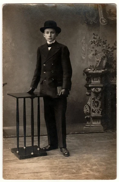 Foto vintage muestra a un niño elegante viste traje negro, guantes negros y sombrero de jugador de bolos. Boy sostiene un pequeño bolsillo (billfold). Fotografía antigua en blanco y negro . — Foto de Stock