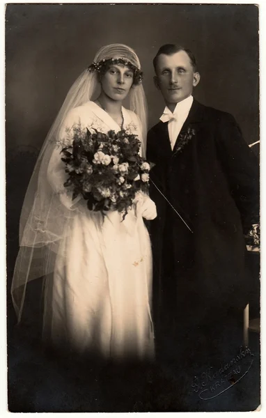 Vintage photo of newlyweds. Bride wears a long veil and holds wedding bouquet. Groom wears black suit and white bow tie. Black & white antique studio portrait. — Stockfoto