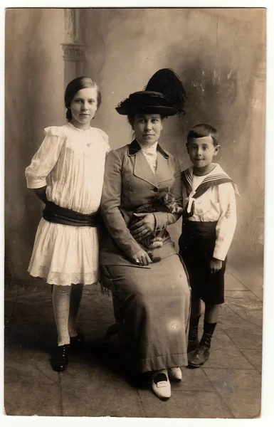 Foto vintage mostra a mãe com seus filhos (menina e menino). Mãe usa um elegante senhoras costum e chapéu de penas, ela segura o gato. Menino usa costum marinheiro e menina usa vestido branco com faixa preta . — Fotografia de Stock