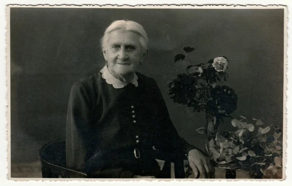 Foto vintage mostra mulher idosa senta-se na poltrona período em um estúdio de fotografia. Na mesa estão flores. Retro preto & fotografia estúdio branco com efeito sépia . — Fotografia de Stock