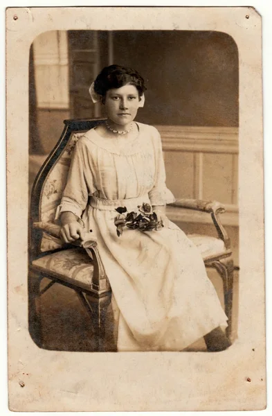 复古照片显示一个年轻女子。她穿了一件白色的裙子。她坐在历史悠久的扶手椅上，捧着玫瑰花。复古黑白工作室摄影. — 图库照片