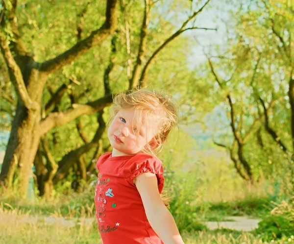 Klein blond meisje zit buiten. Vooraanzicht. Ze draagt een rode blouse groen bomen zijn op achtergrond. — Stockfoto