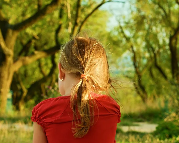 Small blond girl sits outside. Back view. She wears a red blouse, earrings and ponytail.