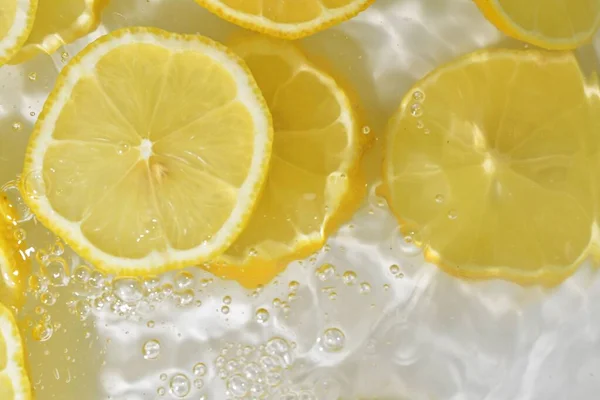 Куски лимона в воде на белом фоне. Лимонный крупный план в жидкости с пузырьками. Кусочки желтого спелого лимона в воде. Макроизображение фруктов в воде — стоковое фото