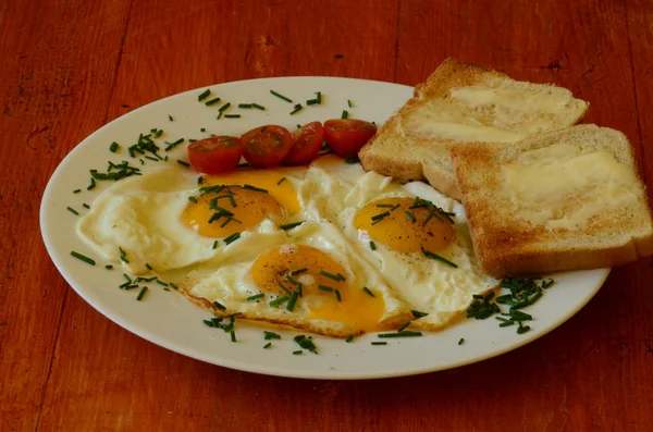 Ηλιόλουστη πλευρά προς τα πάνω τα αυγά με σχοινόπρασο, ντοματίνια και ψητό ψωμί με βούτυρο σε κόκκινο φόντο — Φωτογραφία Αρχείου