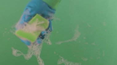 Süngerli lastik eldiven giyen bir el yüzeyi yıkar. Lastik eldiven takan eller süngeri tutar ve yüzeyi temizler. Sünger, krom anahtarın üzerindeki köpüklü deterjanla kaplıdır.