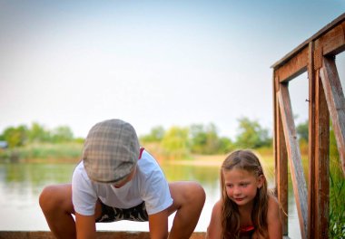 Düz bir şapka takan küçük bir çocuk ve tatlı küçük bir kız gölün kıyısındaki ahşap bir iskelede oturuyorlar. Küçük çocuklar bir göletin kenarındaki iskelede oturuyorlar. Tasasız bir çocukluk ve yaz tatili kavramı.