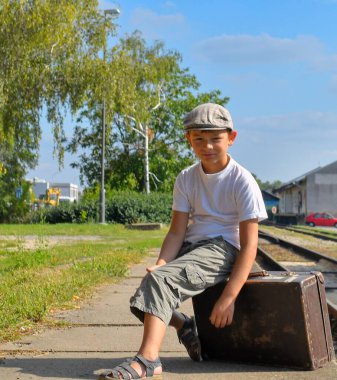 Tren istasyonundaki sevimli çocuk. Gülümseyen küçük bir çocuk eski bir bavula oturur. Seyahat, tatil ve tasasız çocukluk kavramı. Retro ve vintage titreşim