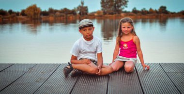 Bir grup şirin çocuk gölün kıyısındaki ahşap iskelede oturuyor. Geleceğe bakan küçük çocuklar. Çocuklar rıhtımda oturuyor. Kardeşler. Farklı yaşlarda iki çocuk okul çağında bir oğlan ve bir anaokulu kızı tahta bir iskelede oturuyorlar. Şey...