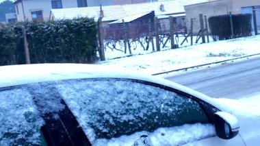 Yakın plan bir araba manzarası beyaz kar tabakası ile tozlanmış, serin bir kış günü işaret eden evler ve çıplak ağaçlar görülebilir.