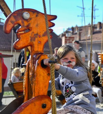 Kedi suratlı bir çocuk garip bir ahşap salıncağın sapını kavrıyor, canlı bir etkinlikte açık havada eğleniyor.