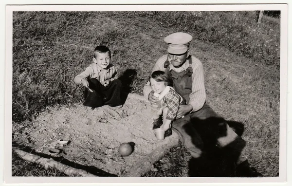 A vintage photo shows grandfather plays with children on sandpit. Antique black & white photo shows photographer shadow. — Stock Photo, Image