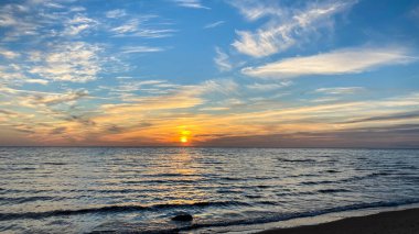 Yaz günbatımında deniz kenarında altın turuncu bir gökyüzü ve bulutların arasında güneş. Martı silueti görünüyor. Plaj kumu deniz suyuyla yıkanır..
