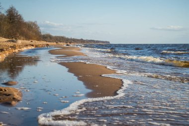 Deniz kıyısındaki güzel sahil, deniz suyu dalgalarıyla yıkanır ıslak kum oluşturur, bu da güzel mavi baharın gökyüzünü yansıtır.