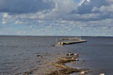 Nehir suyuna batmış ahşap bir patikayı sel basan bir göl manzarası. Su basmış tahta yaya köprüsüne giden kayalık bir yol var. Mavi gökyüzü ve dramatik bulutlar. Bahar manzarası