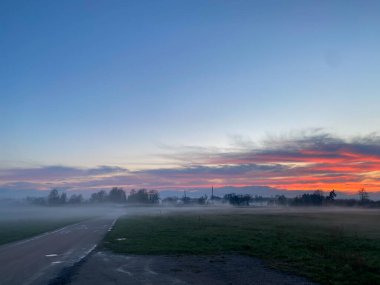 Gün batımında bir otoyola çıkan toprak yol. Kızıl gökyüzü kalın bir sisle kaplı..
