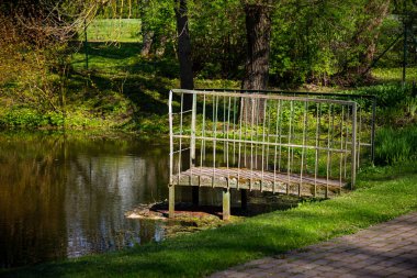 Parktaki göletin üzerinde yeşil metal bir köprü var. Yeşil çimenlerin ve ağaçların yanında..