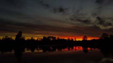 Gölün kenarında güzel, kırmızı bir gün batımı. Kusursuz bir aynanın suyuna yansıyan siyah ağaç siluetleri..