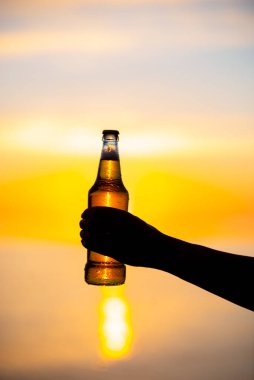 Gün batımında ellerinde bir şişe yaz dönümü temalı bira tutan kadınlar. Bira şişesi güneşten gelen altın ışık, bokeh baloncukları oluşturuyor. Sığ alan derinliği