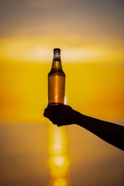 Gün batımında ellerinde bir şişe yaz dönümü temalı bira tutan kadınlar. Bira şişesi güneşten gelen altın ışık, bokeh baloncukları oluşturuyor. Sığ alan derinliği